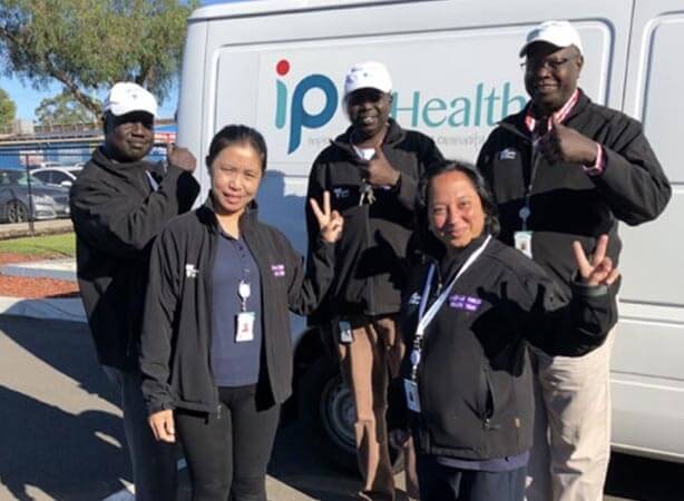 Five bicultural workforce staff stand in front of an IPC Health van and giving a thumbs up or V-sign