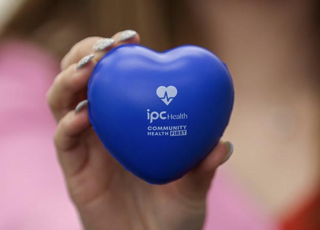 A blue stress ball in the shape of a heart with the IPC Health and Community Health First logos in white text is held up in focus.