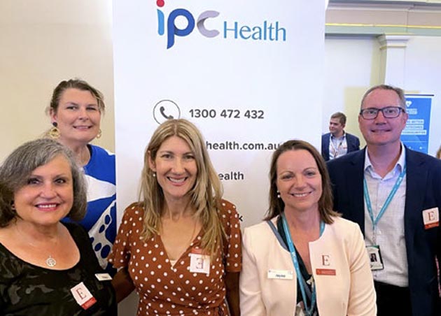 Vilma (Client Services team), Geri (Communications team), Helen (Head to Health team), Jayne Nelson (IPC Health CEO) and Daryl Whitfort (IPC Health Board Chair) stand smiling in front of a white IPC Health branded pull up banners.