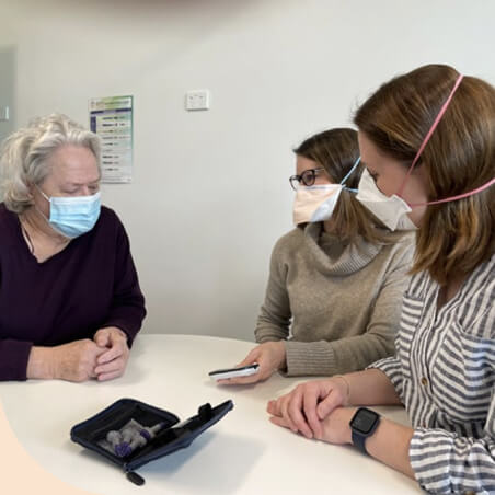 Two IPC Health Diabetes Wellbeing Hub clinicians wearing N95 masks sit at a table with a client. They are talking and explaining the use of diabetes equipment.