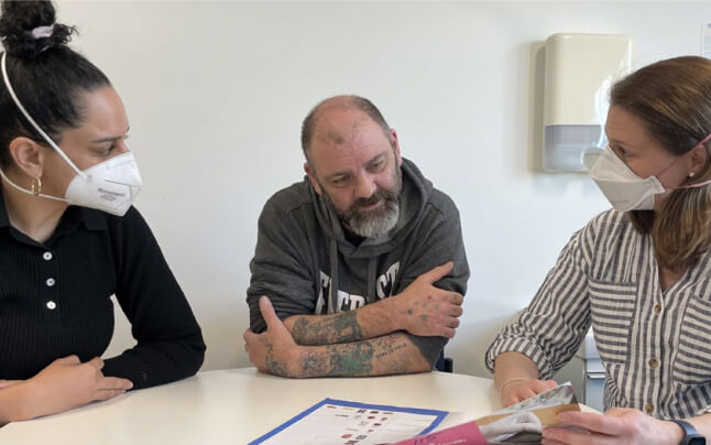 Two IPC Health Diabetes Wellbeing Hub clinicians wearing N95 masks sit at a table with a client. They are talking and sharing information about diabetes.