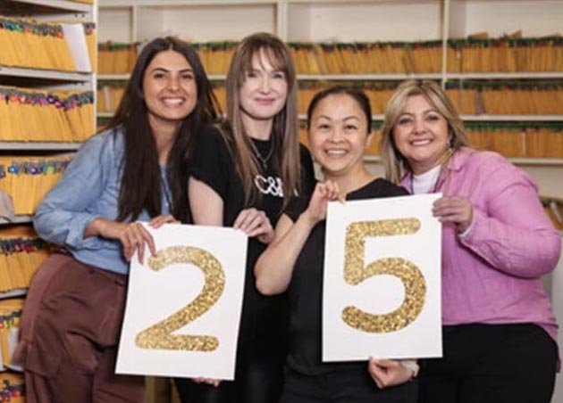 Four Client Services Officers stand together smiling in front of rows of orange folders. They are holding two signs with the number 25 on them.