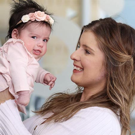 A young mother smiles at her infant baby and holds her up while the baby smiles at the camera.