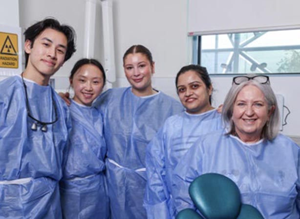 Four students and an IPC Health supervising staff member stand together smiling around a green dental chair. They are wearing blue disposable gowns.