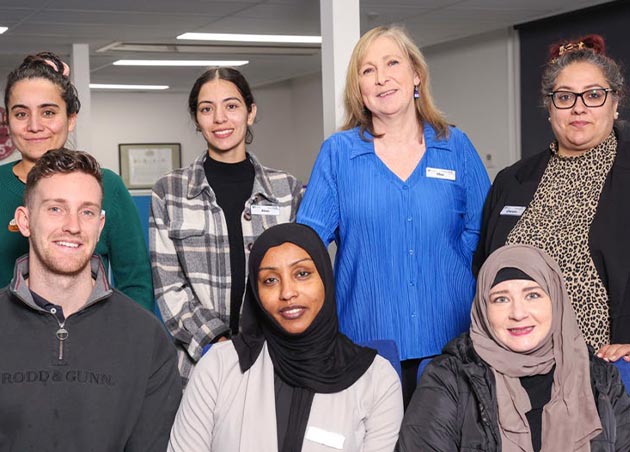 Members of the Homeless and at Risk of Homelessness team are smiling together in an open place office. Three people are sitting on chairs in the front row and four people are standing behind them.