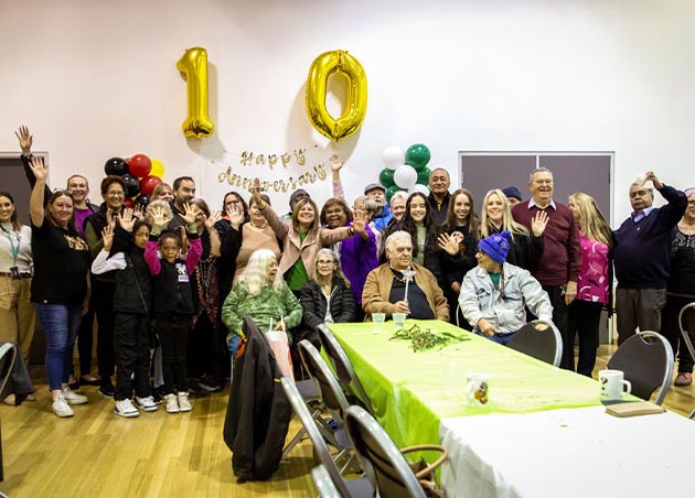 Around 30 people from the IPC
Health Aboriginal and Torres Strait Islander Health team
(including staff and clients) celebrate in front of a banner
that says happy anniversary and two gold balloons in the
shape of the number ten.