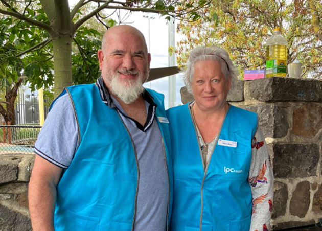 Homeless and at Risk of Homelessness Community Advisory Group volunteers Sam and Corrie stand together smiling while cooking a barbecue. They are both wearing the IPC Health volunteer blue vest.