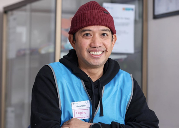 Reception Concierge volunteer Arnold stands smiling in an IPC Health reception area. He is wearing the IPC Health volunteer blue vest.