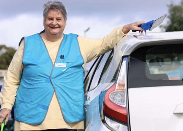 Car wash volunteer Beryl stands smiling next to a white car holding a washing tool. She is wearing the IPC Health volunteer blue vest.